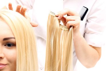 A hairstylist holding blonde hair extensions near a woman's head.