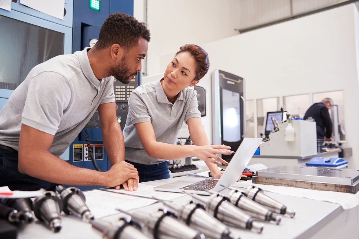 Two engineers discussing work at a laptop in a manufacturing environment.