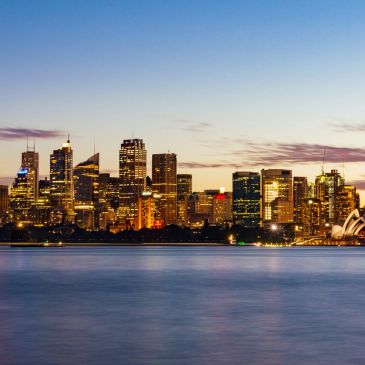City skyline at dusk with illuminated buildings and calm water.