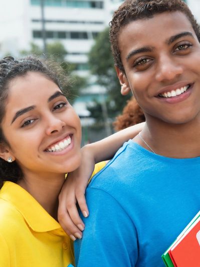 Two young adults smiling outdoors, one with arm around the other.