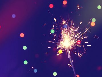 A glowing sparkler shines against a dark, colorful bokeh background.