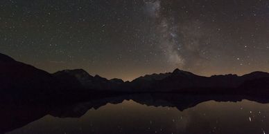 Starry night sky with Milky Way over mountain silhouettes reflected in calm lake.