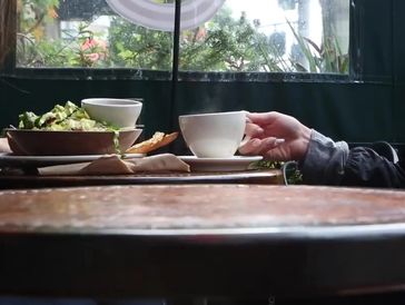 A person holding a cup at a table with food and drink by a window.